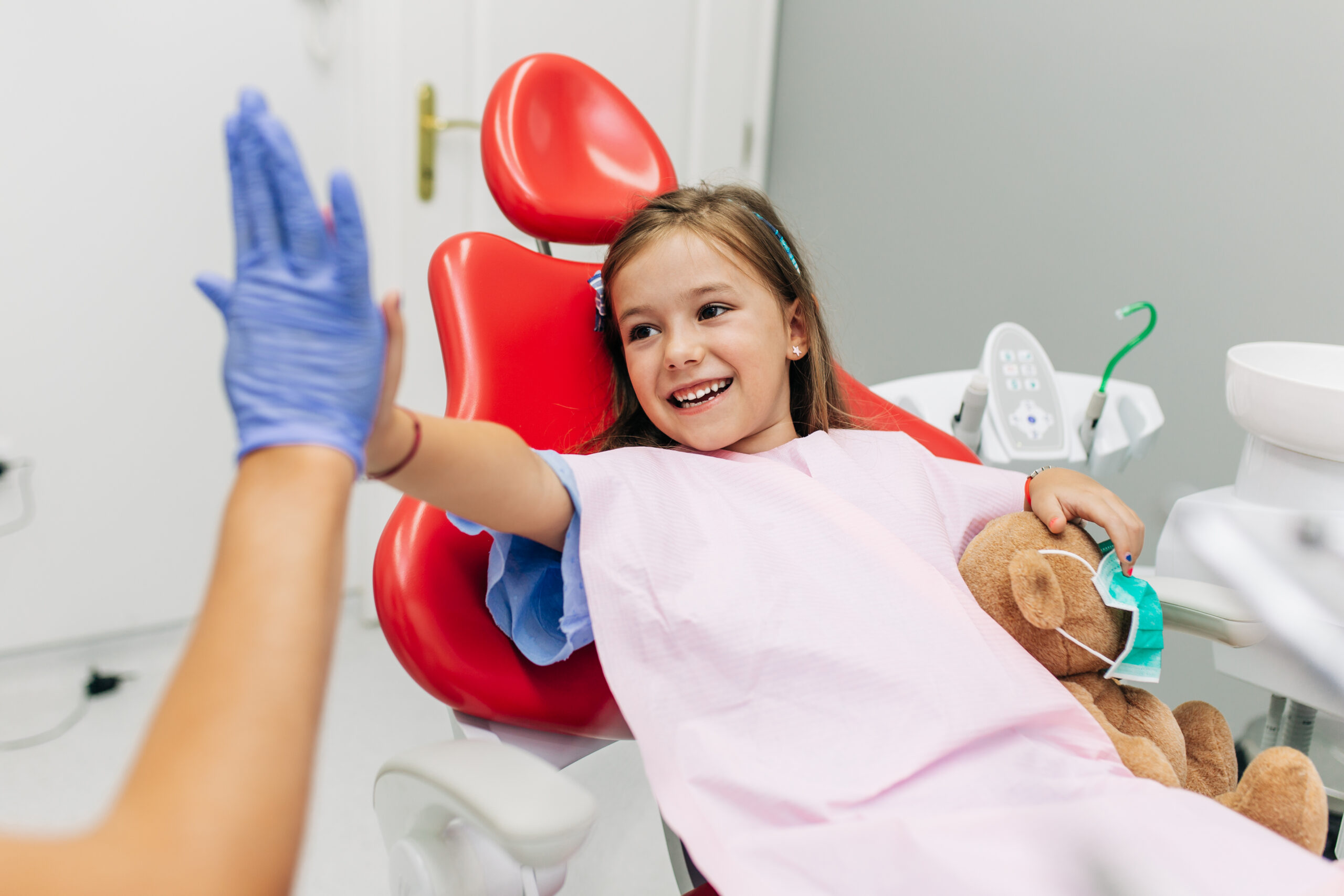 An unseen dentist high fives a little girl laying on a dental chair
